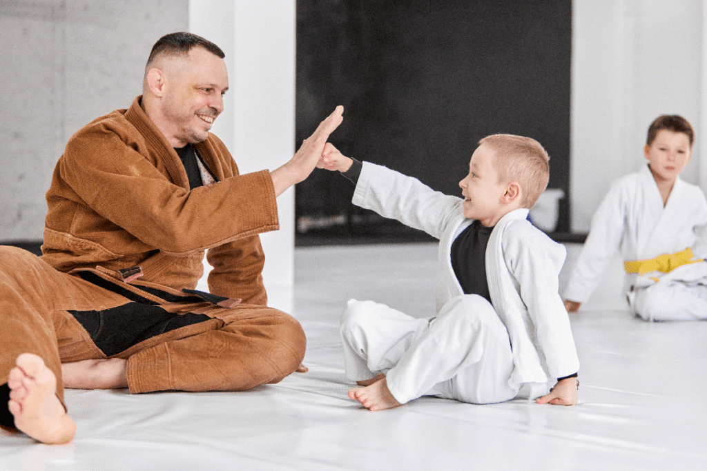 A martial arts instructor high fives a young student while sitting on the mats